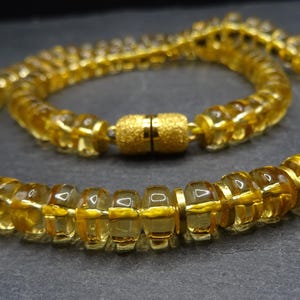 May include: A golden-yellow necklace composed of rounded, segmented beads. The beads are strung together, forming a continuous loop, and secured with a gold-colored clasp. The necklace is displayed against a dark background, highlighting its color and design.