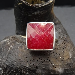 May include: A silver ring featuring a square-cut, faceted ruby gemstone. The bright red stone is set in a minimalist design. The ring is displayed on a dark, textured surface, accentuating the gemstone's colour and sparkle.