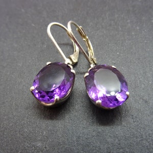 May include: A pair of leverback earrings featuring large, oval-cut, deep purple gemstones. The gemstones are set in a silver-colored metal. The earrings are displayed against a dark gray background.