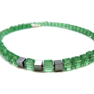 May include: A green beaded necklace with square-cut beads. The necklace features a series of small, metallic gray cube-shaped beads interspersed among the green beads. The necklace is displayed against a white background.