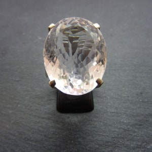 May include: A large, faceted, oval-cut gemstone ring. The clear, light-colored stone is set on a gold-colored band. The ring is displayed on a black stand against a dark background.