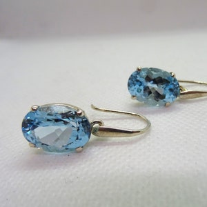 May include: A pair of earrings featuring oval-cut, light blue gemstones. The gemstones are set in a gold-colored metal with a hook closure. The earrings are displayed against a white background.