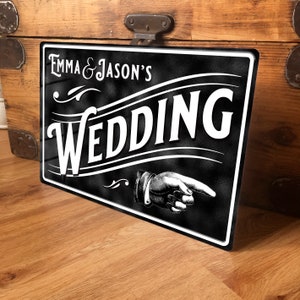 May include: A black and white wedding sign, featuring the text "Emma & Jason's Wedding". The sign has a vintage design with a pointing hand illustration. The rectangular sign has a white border.