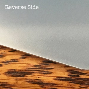 Pu&ograve; includere: Primo piano del retro di un oggetto rettangolare liscio di colore grigio chiaro. L'oggetto &egrave; appoggiato su una superficie di legno marrone con un motivo a strisce.