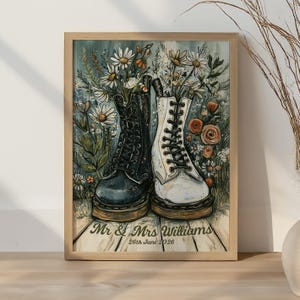 Op de afbeelding: Een ingelijste wandkunstprint met een zwarte en een witte veterlaars gevuld met witte madeliefjes en oranje bloemen. De tekst luidt Mr & Mrs Williams 26th June 2026. De laarzen staan op een houten ondergrond met een lichtblauwe achtergrond.