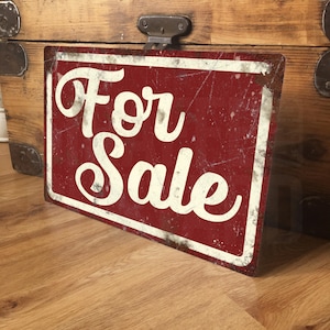 May include: A rectangular metal sign with a distressed, weathered finish featuring a deep red background and a white border. The words For Sale are written in a large, white, cursive script font across the centre of the sign surface.