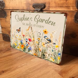 May include: A metal sign with a rustic, weathered look featuring a floral design with a bluebird and a butterfly. The sign reads "Sophie's Garden Sit, Relax & Enjoy".