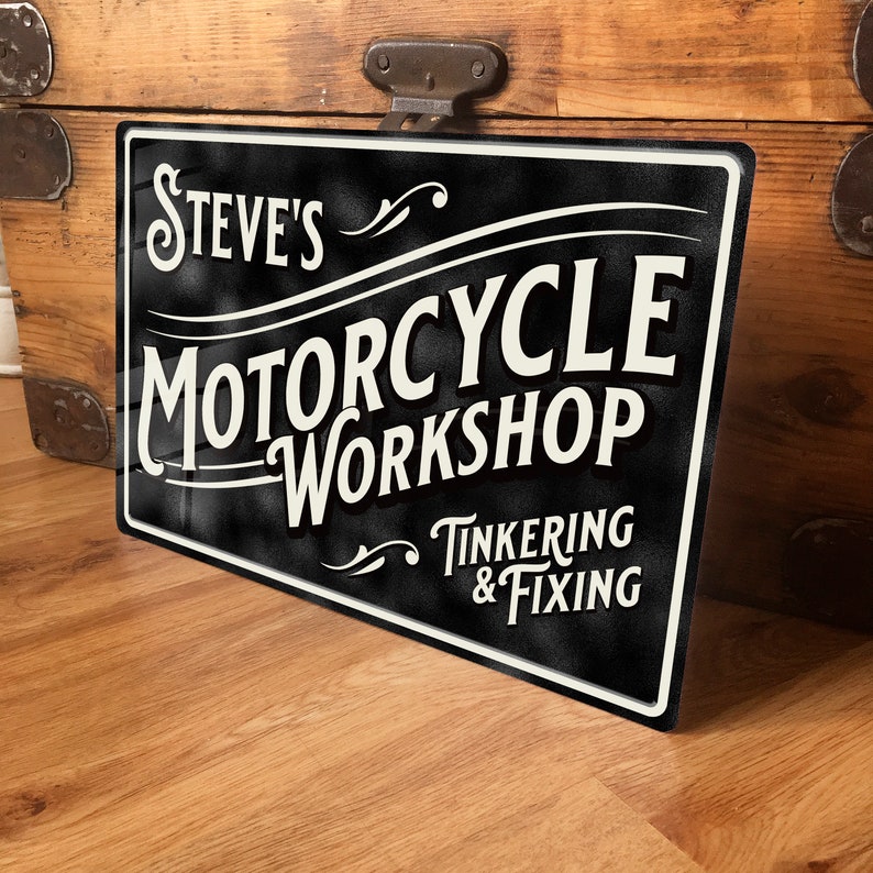 Puede incluir: Letrero met&aacute;lico en blanco y negro que dice "Steve's Motorcycle Workshop" con las palabras "Tinkering & Fixing" debajo. El letrero est&aacute; sobre una superficie de madera.
