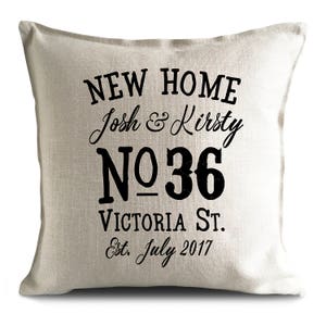 May include: A beige throw pillow with black text that reads "NEW HOME Josh & Kirsty NO 36 VICTORIA ST. Est. July 2017".