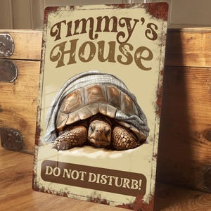 May include: A rectangular metal sign with a distressed, vintage appearance. It features a detailed illustration of a tortoise under a blanket, with the text "Timmy's House" above and "DO NOT DISTURB!" below. The sign has a brown and beige colour scheme.