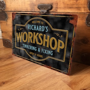 May include: A rectangular metal sign with a distressed black background and gold lettering. The sign reads "Welcome to Richard's Workshop, Tinkering & Fixing, Open 24 Hours." The sign has a rusty border and is mounted on a wooden surface.