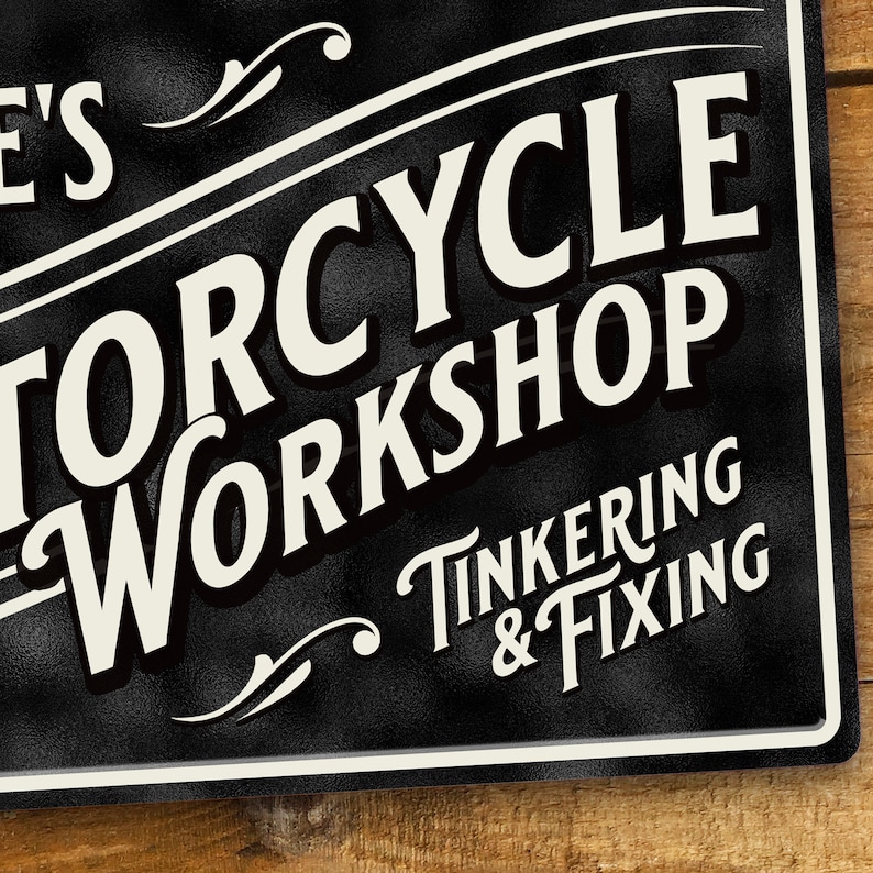 Puede incluir: Letrero met&aacute;lico negro con texto blanco que dice "MOTORCYCLE WORKSHOP" y "TINKERING & FIXING". El letrero tiene un borde blanco y est&aacute; sobre un fondo de madera.