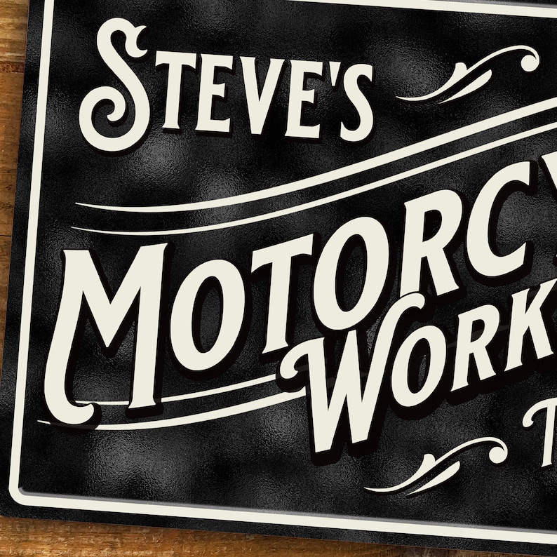 Puede incluir: Letrero de metal negro con el texto "Steve's Motorcycle Workshop" en un estilo vintage. El letrero tiene una superficie texturizada y letras blancas con un borde decorativo.
