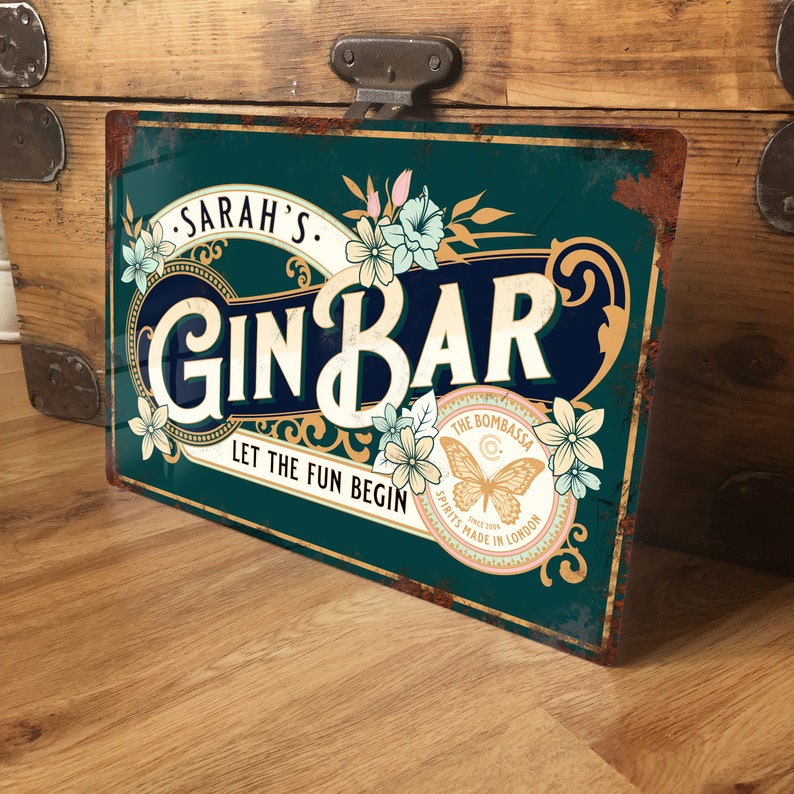 Pu&ograve; includere: Un cartello in metallo blu-verde con bordo dorato e dettagli floreali. Il cartello recita "SARAH'S GIN BAR LET THE FUN BEGIN" con un logo di farfalla dorato e il testo "THE BOMBASSA SPIRITS MADE IN LONDON".
