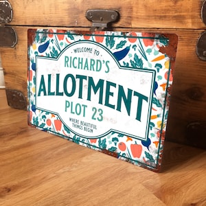 May include: A white metal sign with a green border and a rusty texture. The sign says "Welcome to Richard's Allotment Plot 23 Where Beautiful Things Begin" and is decorated with colourful vegetables and flowers.
