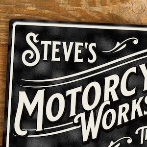 Puede incluir: Letrero de metal negro con letras blancas que dice "Steve's Motorcycle Works". El letrero tiene una superficie texturizada y un borde decorativo. El texto est&aacute; en una fuente vintage y estilizada.