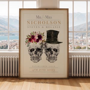 Op de afbeelding: Een rechthoekige print met de tekst MR & MRS NICHOLSON, STEVEN & MELANIE en ESTABLISHED SINCE 4TH JUNE 2023. Het ontwerp toont twee menselijke schedels, één versierd met een bloemenkrans van rode en roze bloemen en één met een zwarte hoge hoed.