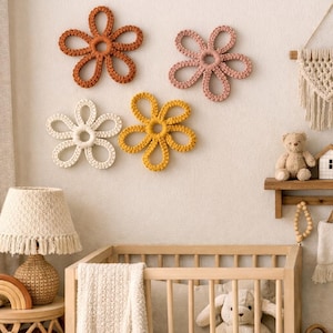 Könnte beinhalten: Vier gehäkelte Blumen-Wanddekorationen in Orange-, Rosa-, Gelb- und Weißtönen sind an einer neutralen Wand angebracht. Ein Holzbettchen, eine Lampe mit Fransen-Schirm und ein kleines Regal mit einem Teddybären und einem Haus sind ebenfalls zu sehen.