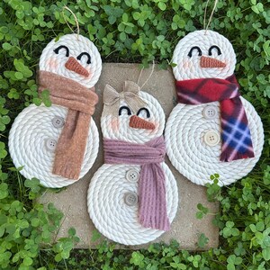 May include: Three handmade snowman ornaments. Each snowman is crafted from coiled white rope, featuring button accents, fabric scarves in various colors, and a carrot nose. The snowmen have black eyes and are set against a backdrop of green foliage.