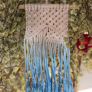 May include: A macrame wall hanging with a natural wood dowel and a cream-colored woven top. The bottom fringe transitions from cream to a light blue, creating an ombre effect. The piece is suspended by a cream-colored cord.