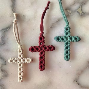 May include: Three macrame crosses in beige, burgundy, and teal. Each cross has a loop for hanging. The crosses are made of knotted cord, with a textured appearance. The background is a light-colored, marbled surface.