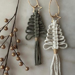 Puede incluir: Dos adornos navideños de macramé con forma de árbol. Uno es verde oliva, el otro blanco roto. Cada uno tiene una estrella de madera en la parte superior y una borla en la parte inferior, colgados de cuerdas. Una rama con bayas doradas brillantes está en el fondo.