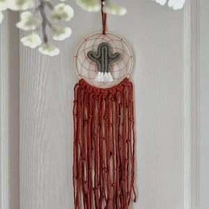 May include: A handmade dreamcatcher featuring a gray cactus design in the center. The dreamcatcher has a white circular frame with a webbed interior. Long, rust-colored knotted cords hang below, creating a bohemian aesthetic.