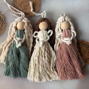 Pode incluir: Três bonecas de macramé feitas à mão com cabeças de contas de madeira e cabelo comprido de fio. As bonecas são em tons de verde, creme e rosa empoeirado, com detalhes intrincados de macramé em seus corpos. Cada boneca tem um laço para pendurar.