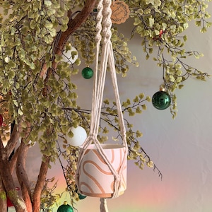 Może przedstawiać: Makrama do wieszania roślin w kolorze beżowym i kremowym. Wisi na drewnianym pierścieniu i trzyma różową ceramiczną doniczkę z falistym, abstrakcyjnym wzorem. Tło obejmuje drzewo z zielonymi liśćmi i ozdobami świątecznymi.