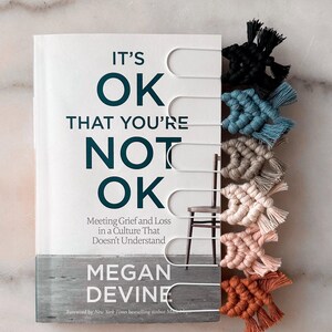 May include: A book titled "IT'S OK THAT YOU'RE NOT OK" with a white cover and teal lettering. The book is accompanied by a set of handmade macrame bookmarks in various colors, including black, teal, and beige.
