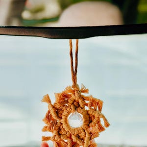 May include: A tan-coloured macrame car charm in a starburst design. The charm is made of knotted cord and features a clear, faceted crystal in the centre. The charm hangs from two twisted cords.