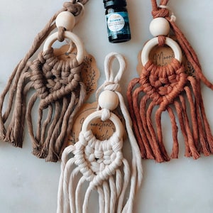 Puede incluir: Tres ambientadores de coche de macramé en tonos marrón, crema y óxido, cada uno con un anillo y una cuenta de madera. También hay una pequeña botella de aceite esencial con tapa negra. Estilo bohemio.