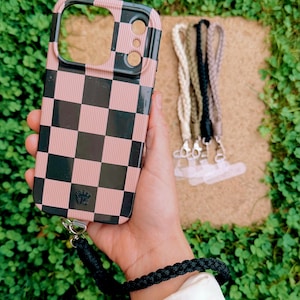 May include: A pink and black checkered phone case with a black border. The case has a wrist strap attached. Several braided phone lanyards in cream, black, and brown are also visible.
