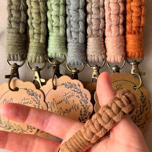 Op de afbeelding: Een verzameling macramé sleutelhangers in verschillende kleuren, waaronder olijfgroen, grijs en perzik. Elke sleutelhanger heeft een geweven ontwerp en een metalen sluiting. De sleutelhangers worden gepresenteerd met decoratieve labels met de tekst "Moonstone Marvels".