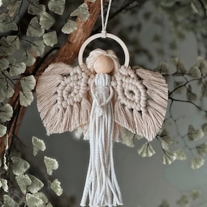 May include: A macrame angel ornament with a wooden head and ring, beige wings, and white fringe. The angel is suspended from a branch, showcasing a handcrafted, bohemian style. The background features green foliage.