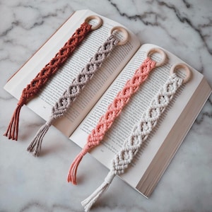 May include: Four macrame bookmarks in shades of rust, gray, peach, and white, each with a wooden ring and tassel. The bookmarks are laid across an open book, showcasing their intricate woven design.