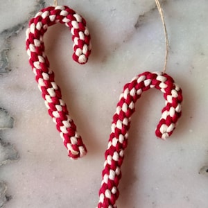 Könnte beinhalten: Zwei handgefertigte Weihnachtsornamente in Form von Zuckerstangen. Die Zuckerstangen sind rot und weiß, aus geflochtenem Seil gefertigt und haben eine Schnurschlaufe zum Aufhängen. Auf einer Marmoroberfläche präsentiert.