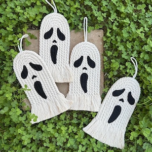 May include: Four white macrame ghost decorations with black facial features and a looped string for hanging. The ghosts have a fringed bottom and are arranged on a bed of green clover.