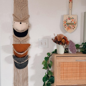 Op de afbeelding: Een beige macramé hoedenorganizer hangt aan een witte muur en bevat vier baseballpetten in verschillende kleuren. Een decoratieve schop met bloemmotieven is in de buurt gemonteerd. Een houten kast met een geweven voorkant en een plant maken de scène compleet.