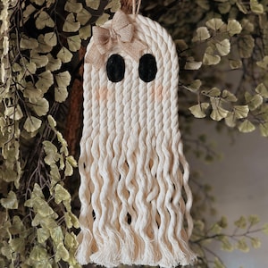 Könnte beinhalten: Handgefertigte Makramee-Geisterdekoration mit einer Schleife aus Jute und schwarzen Filzaugen. Der Geist besteht aus cremefarbenem Seil und hat einen Fransenboden. Eine dekorative Hängedekoration für Halloween.