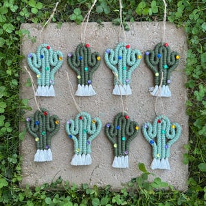 May include: Eight macrame cactus ornaments in shades of green, adorned with colorful bead accents. Each cactus has a white fringe base and a twine loop for hanging. The ornaments are arranged on a stone surface surrounded by green foliage.