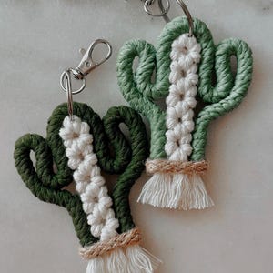 Pode incluir: Dois chaveiros de cacto em macramé. O design do cacto apresenta corda verde com um centro branco e um detalhe de juta natural na base. Cada chaveiro tem um fecho prateado.
