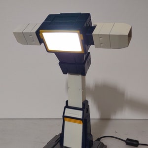 May include: A modern desk lamp with a unique design. The lamp features a rectangular light source, flanked by two off-white panels. The base and supporting structure are a mix of gray, white, and navy blue. The lamp is plugged into a power outlet.