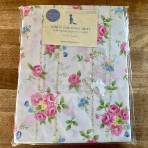 May include: A white crib fitted sheet featuring a floral pattern of pink roses, green leaves, and small blue flowers with vertical yellow stripes. The packaging includes a label reading Ashley Crib Fitted Sheet and Pottery Barn Kids.