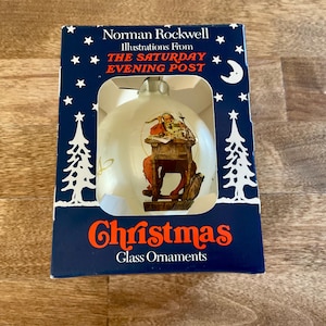 May include: A white glass Christmas ornament featuring a Norman Rockwell illustration of a person at a desk, packaged in a dark blue box with white stars, trees, and the text Norman Rockwell Illustrations From THE SATURDAY EVENING POST Christmas.