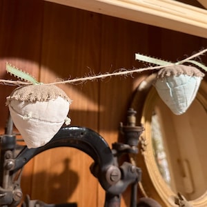 May include: A handmade garland featuring fabric acorn ornaments. Each acorn has a white body, a burlap cap, and a green ribbon stem. The garland is strung on twine, creating a rustic and charming decorative piece.