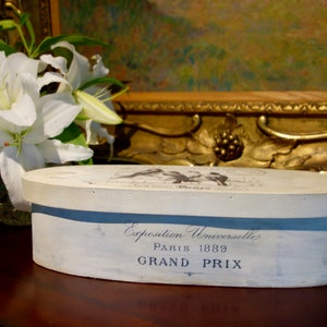 May include: An oval wooden box with a distressed white finish. The side features dark blue text reading Exposition Universelle PARIS 1889 GRAND PRIX. The lid displays a graphic of birds and additional text. The box has a smooth, flat top surface.