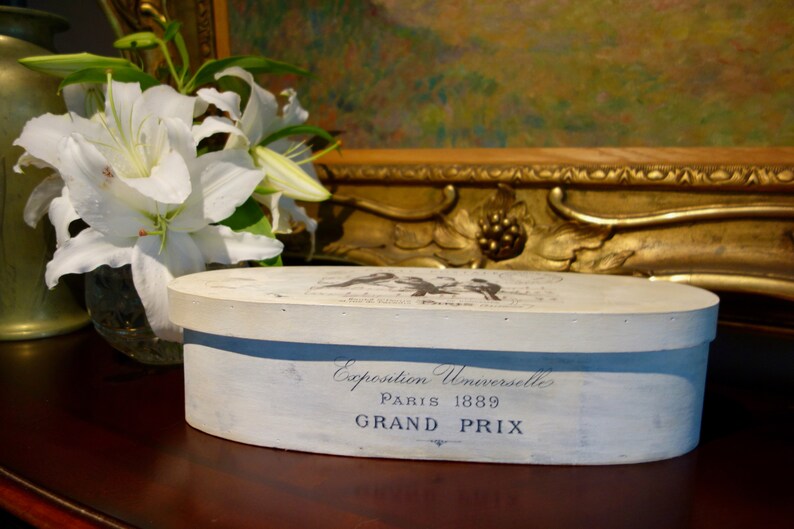 May include: An oval wooden box with a distressed white finish. The side features dark blue text reading Exposition Universelle PARIS 1889 GRAND PRIX. The lid displays a graphic of birds and additional text. The box has a smooth, flat top surface.