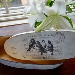 May include: An oval wooden box with a distressed white finish. The lid features a vintage postcard graphic with four birds perched on musical notes, the text "CARTE POSTALE," and a "TOUR EIFFEL" stamp. The interior is natural wood.