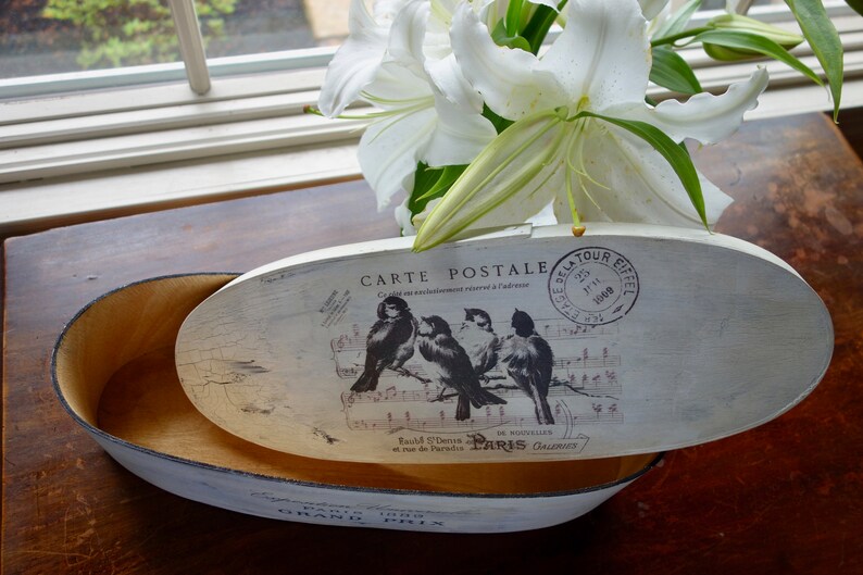 May include: An oval wooden box with a distressed white finish. The lid features a vintage postcard graphic with four birds perched on musical notes, the text "CARTE POSTALE," and a "TOUR EIFFEL" stamp. The interior is natural wood.